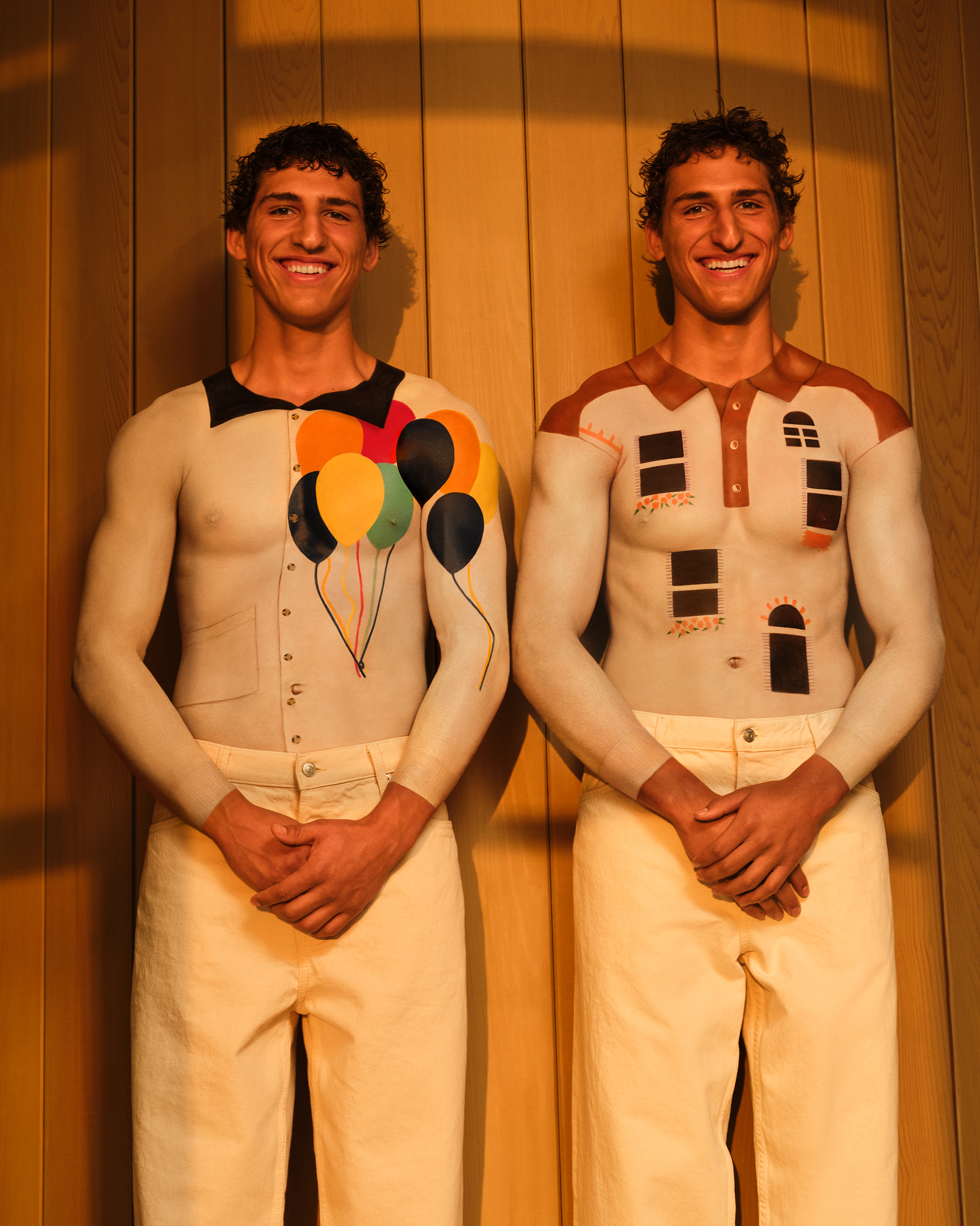 Twin brothers with white body painted tops smiling and looking at the camera against a wooden wall